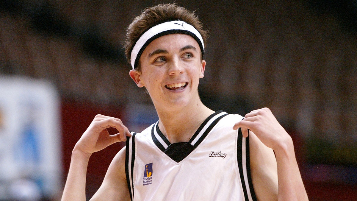 Frankie Muniz in einem Basketballtrikot und passendem Schweißband hält die Ärmel seines Trikots hoch und lächelt