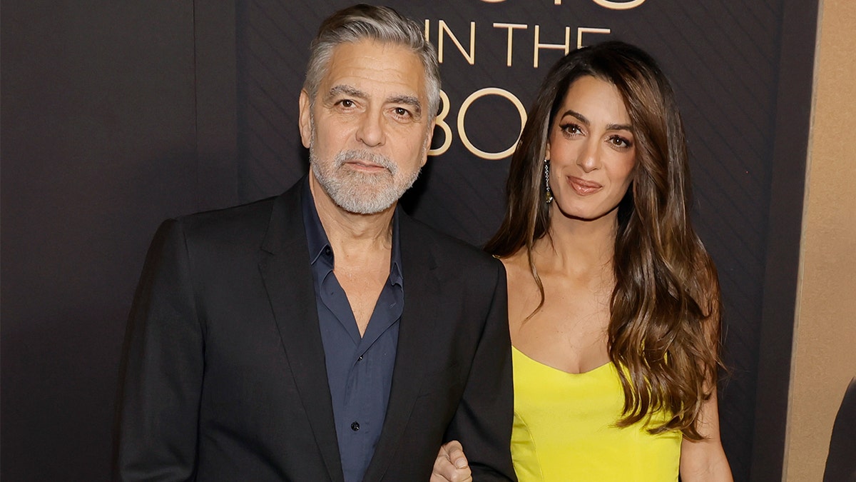 George Clooney in schwarzem Blazer und blauem Button-Down steht neben Frau Amal in einem gelben Kleid