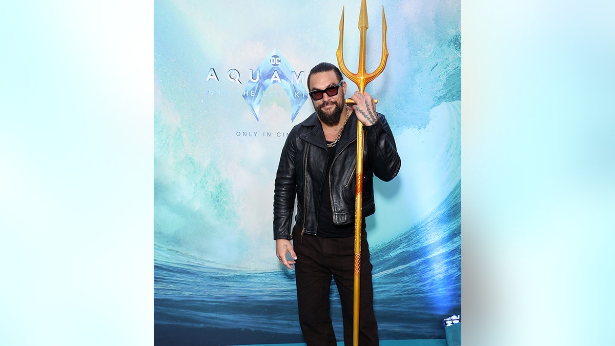 Jason Momoa in einem schwarzen Lederoutfit hält den Dreizack auf "Aquaman" roter Teppich