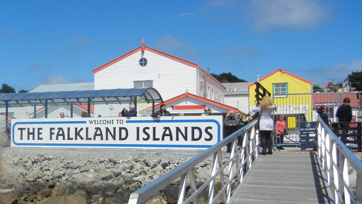 Schild der Falklandinseln vor ein paar Gebäuden, Menschen auf der Promenade