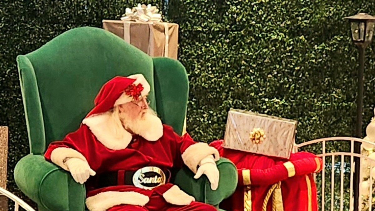 Der Weihnachtsmann sitzt in einem großen grünen Stuhl und schaut nach rechts, vor Büschen, neben Weihnachtsdekoration