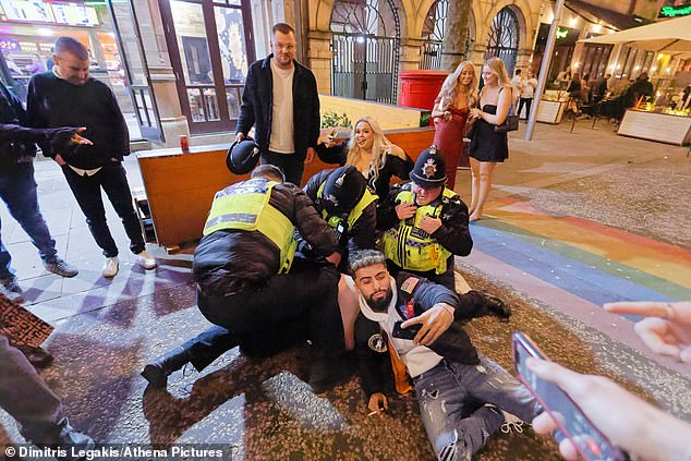 In this shot, one of the men is now on the ground and taking a selfie in front of the officers, while another holds a policemen's hat and a third poses for the camera in front of a large group of spectators