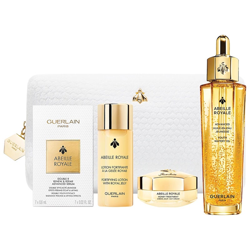 Guerlain Abeille Youth Watery Oil Set: Vier verschiedene goldfarbene Hautpflegeprodukte von Guerlain neben einer weißen Kulturtasche auf weißem Hintergrund
