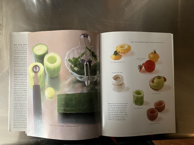 Martha Stewart book, open on a metal counter. Pages show a scooped-out cucumber and other scooped-out fruits and vegetables, including a grape.