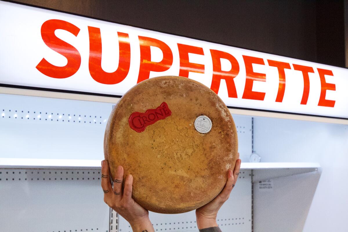 Eine Hand hält einen Laib Käse vor einen Kühlschrank mit der Aufschrift „ "SUPERETTE" bei DTLA Cheese Superette.