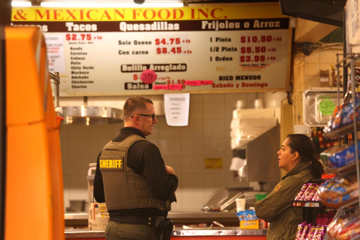 Ein Mann (links) mit Brille und olivgrüner Weste und dem Wort „Sheriff“ spricht mit einer Frau in einem Geschäft 