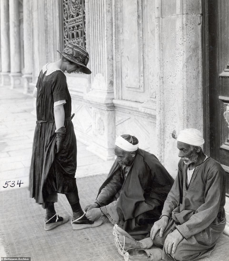 A western tourist waits outside a mosque as an attendant ties slippers to her shoes so she doesn't need to take them off. Today tourists traditionally remove their shoes when entering a mosque instead of wearing overshoes