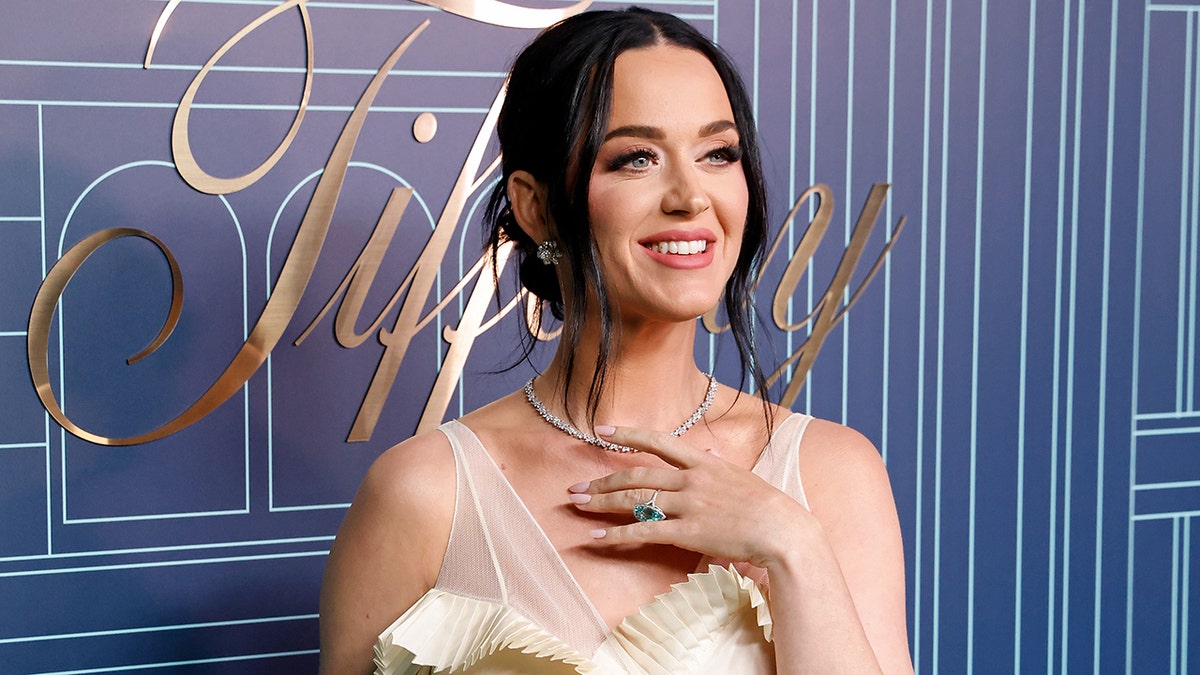 Katy Perry blickt in einem champagnerfarbenen Kleid mit der Hand auf der Brust von der Kamera weg