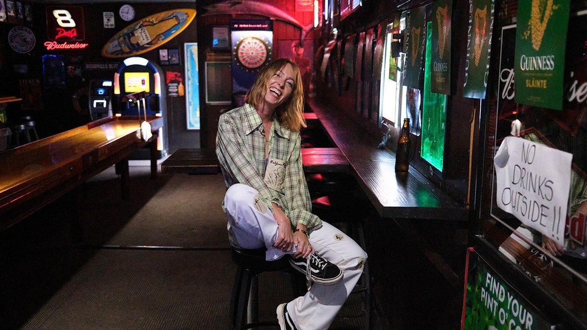India Oxenberh trägt ein kariertes Hemd und eine weiße Hose, sitzt lächelnd in einer Bar