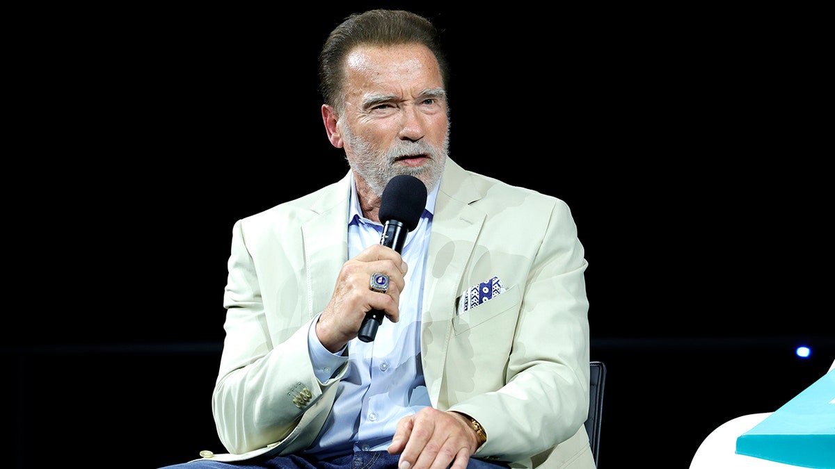 Arnold Schwarzenegger im hellblauen Hemd und cremefarbenen Anzug spricht auf der Bühne ins Mikrofon