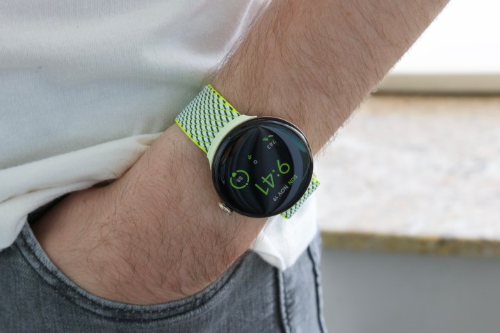Jemand trägt die Google Pixel Watch 2 mit einem gelb/grünen Stoffarmband.