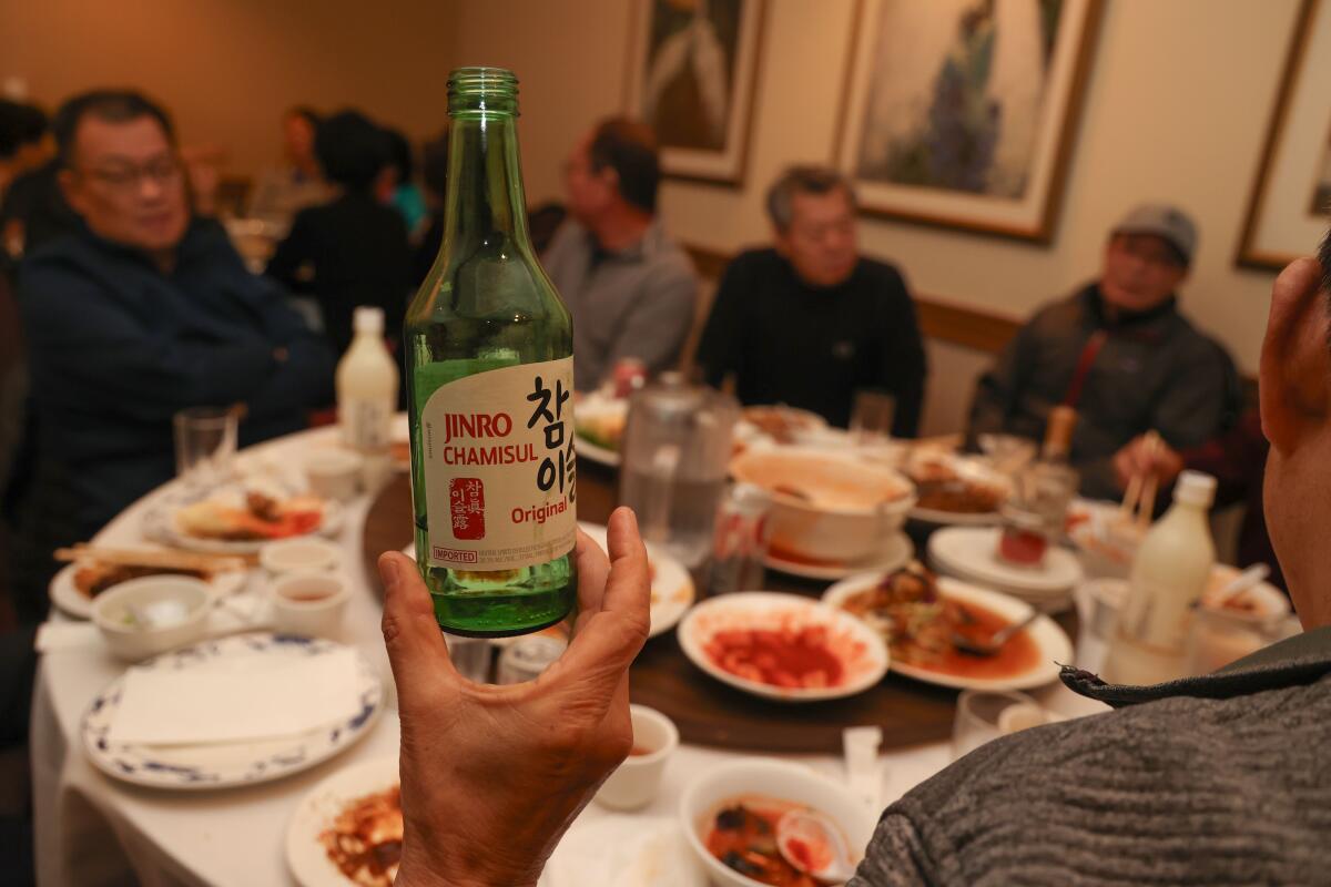 Eine Hand hält eine grüne Glasgetränkeflasche in einem Speisesaal voller Menschen an runden Tischen hoch.