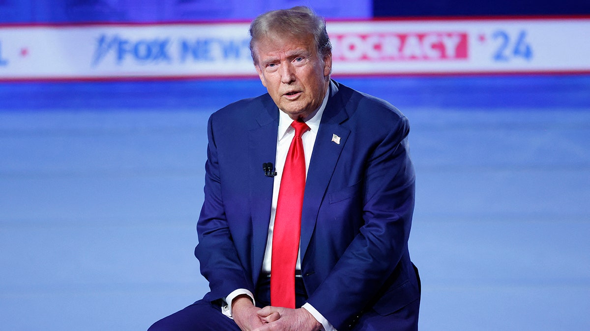 Trump redet, sitzt auf einem Hocker auf der Bühne, verschränkt die Hände auf dem Schoß und trägt einen dunkelblauen Anzug mit einer knallroten Krawatte