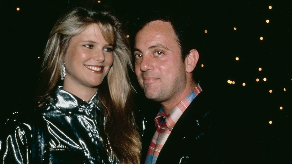 Christie Brinkley lächelt und schaut leicht herab auf Billy Joel in einer schwarzen Jacke und einem bedruckten Hemd mit Kragen