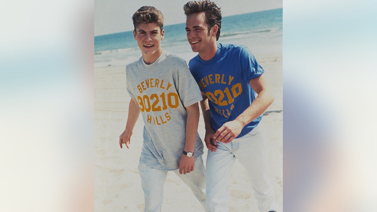 Brian Austin Green in einem grauen Beverly Hills 90210-Shirt und Luke Perry in einem blauen Beverly Hills 90210-Shirt gehen gemeinsam am Strand spazieren