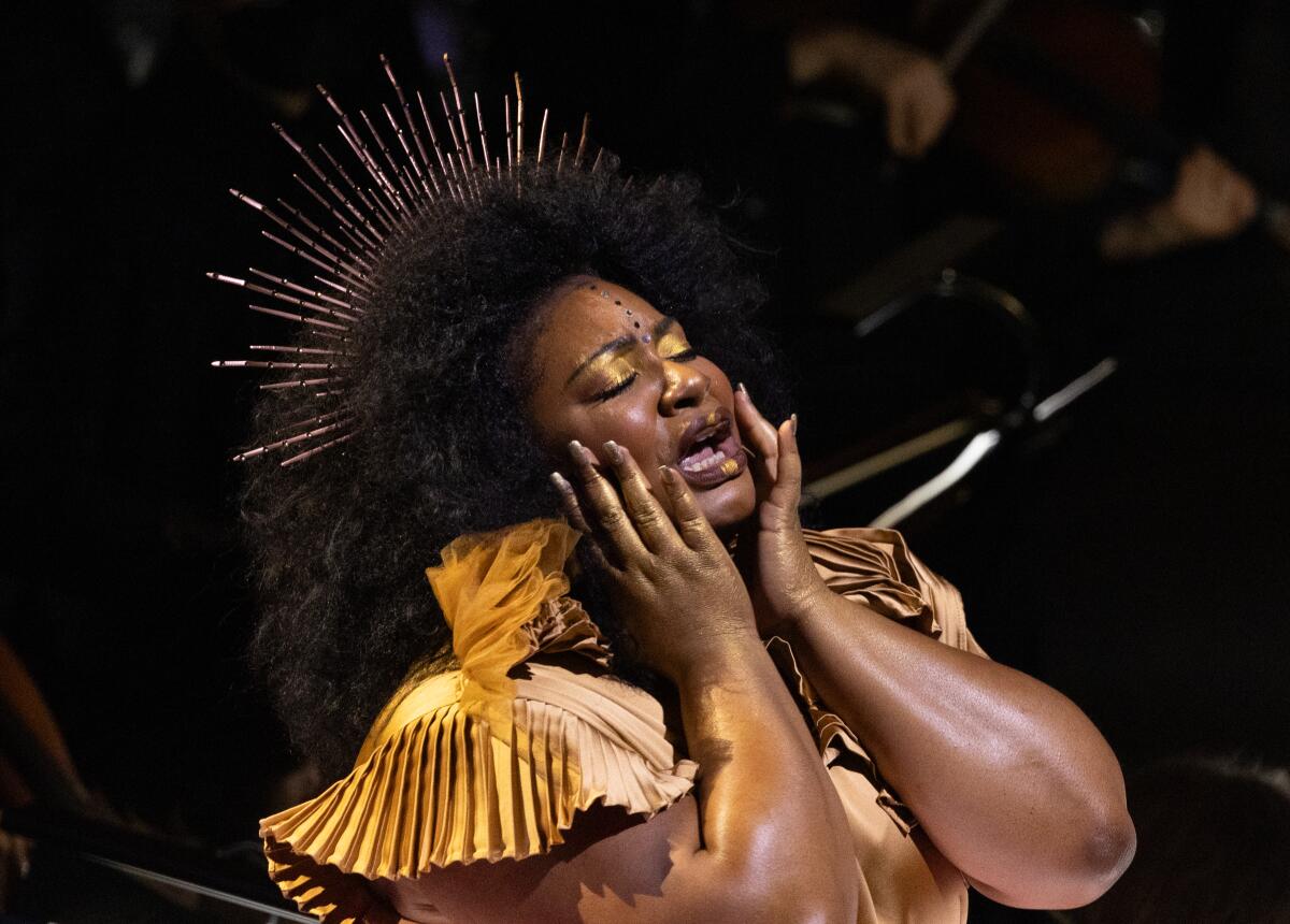 Raehann Bryce Davis, eine schwarze Frau in einem goldenen Kleid mit einer goldenen Krone, schließt die Augen und legt ihre Hände auf ihre Wangen.