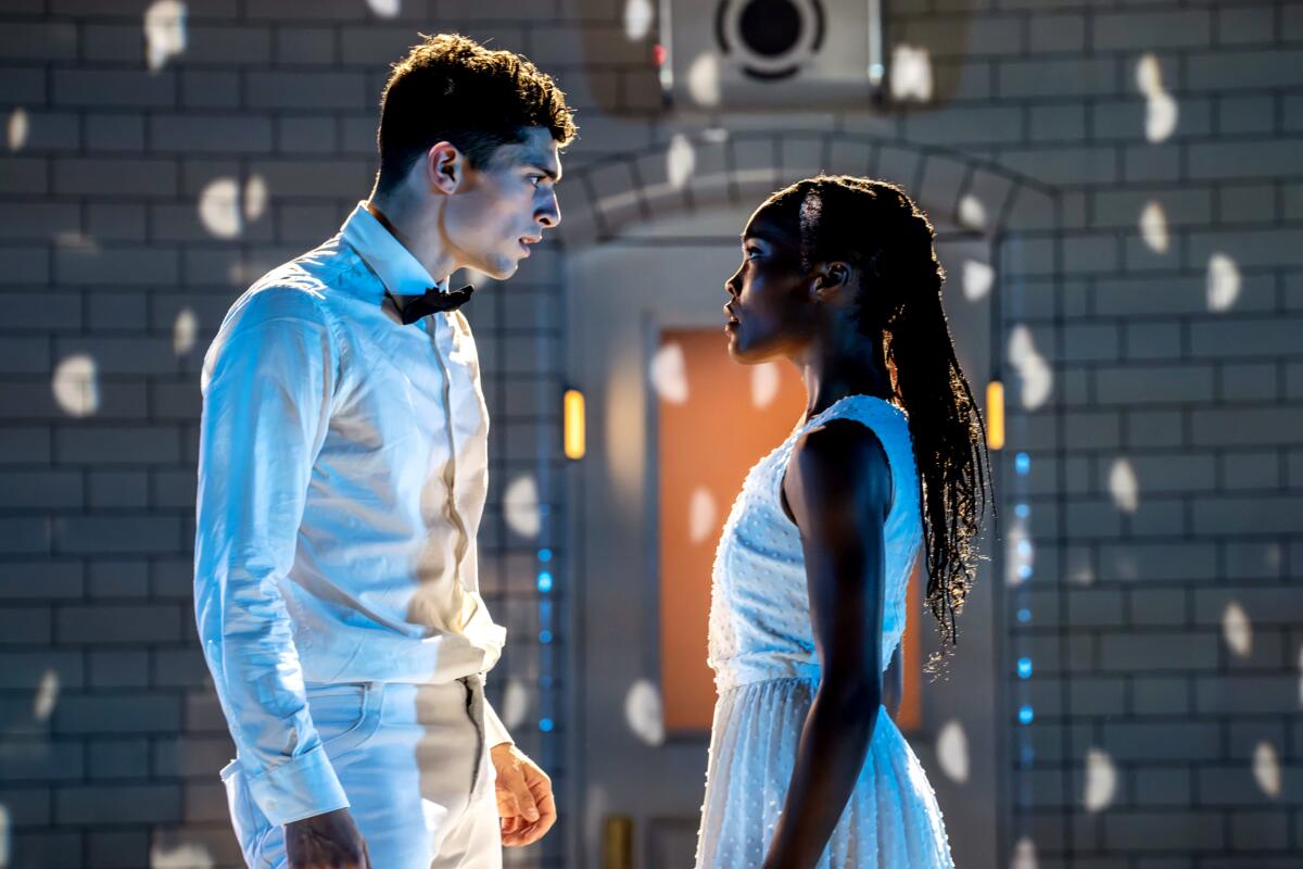 Zwei weiß gekleidete Tänzer – ein weißer Mann und eine schwarze Frau – stehen sich auf einer dramatisch beleuchteten Bühne gegenüber.