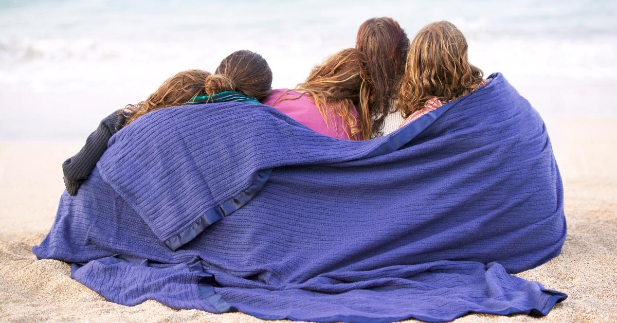 Rückansicht einer Gruppe von Freunden, die zusammen in eine Decke gehüllt am Strand sitzen – Stockfoto