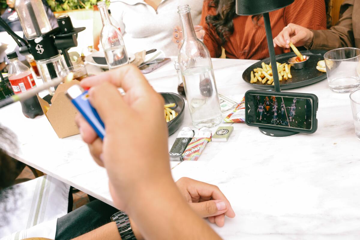 Eine Nahaufnahme einer Hand, die einen Joint anzündet, neben einem Smartphone, das auf einem Tisch liegt und ein Fußballspiel überträgt
