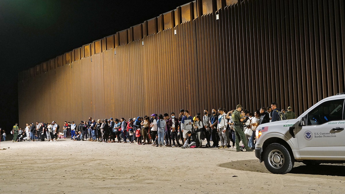 Migranten stehen in einer Schlange entlang der Grenzmauer