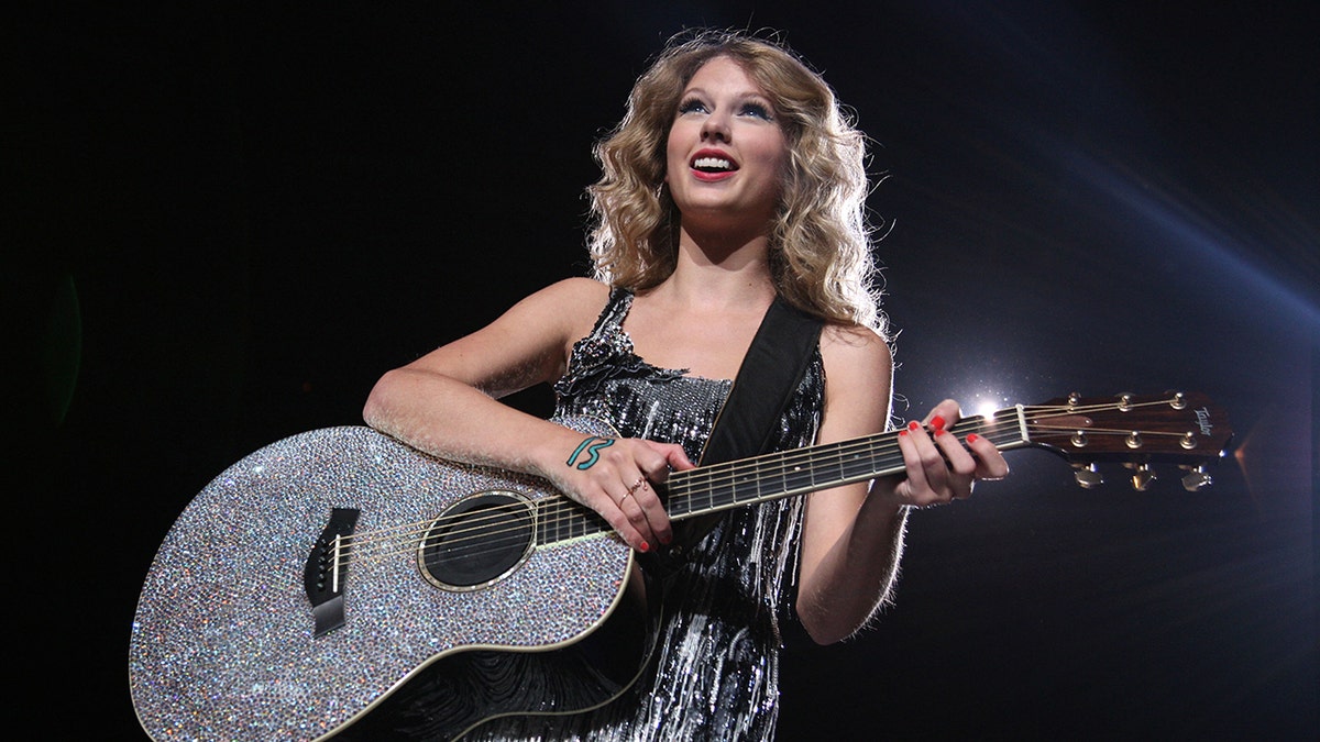 Taylor Swift in einem glitzernden Kleid spielt auf einer juwelenbesetzten Gitarre, auf deren Hand die Nummer 13 steht