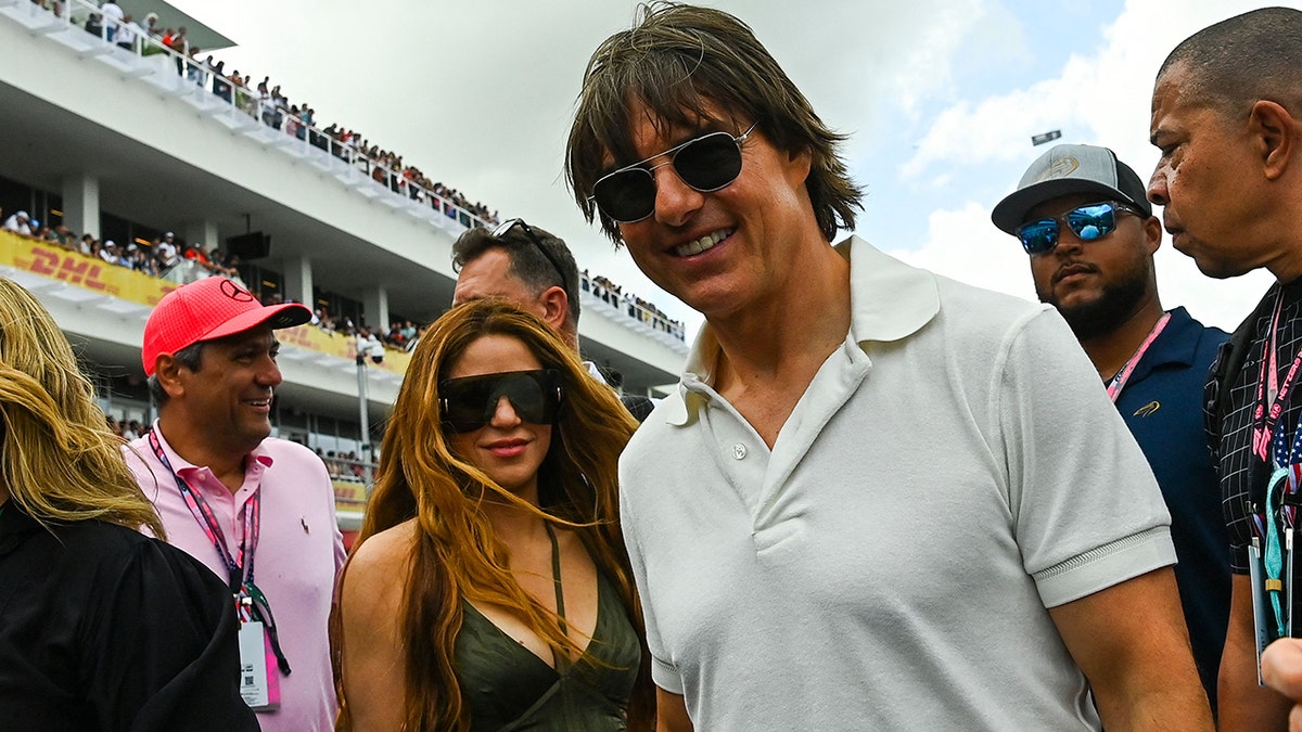 Shakira in einem schwarzen Tanktop mit zwei Schnüren in der Mitte um den Hals und einer riesigen Brille lächelt sanft für ein Foto mit Tom Cruise in einem weißen Poloshirt beim Grand Prix