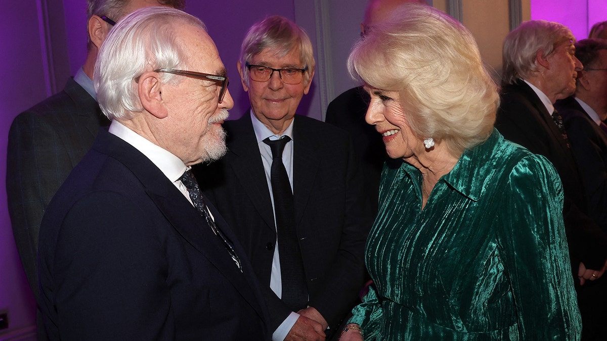 Brian Cox schüttelt Königin Camilla die Hand, die ein samtgrünes Kleid trägt