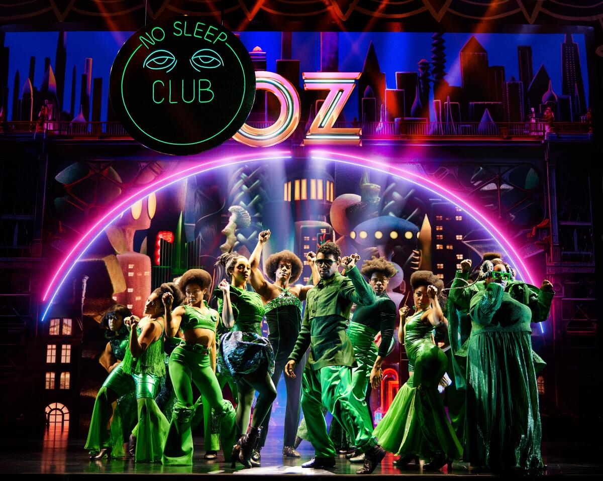 In Grün gekleidete Schauspieler unter einem Neonbogen mit dem Wort „Oz“ an der Spitze