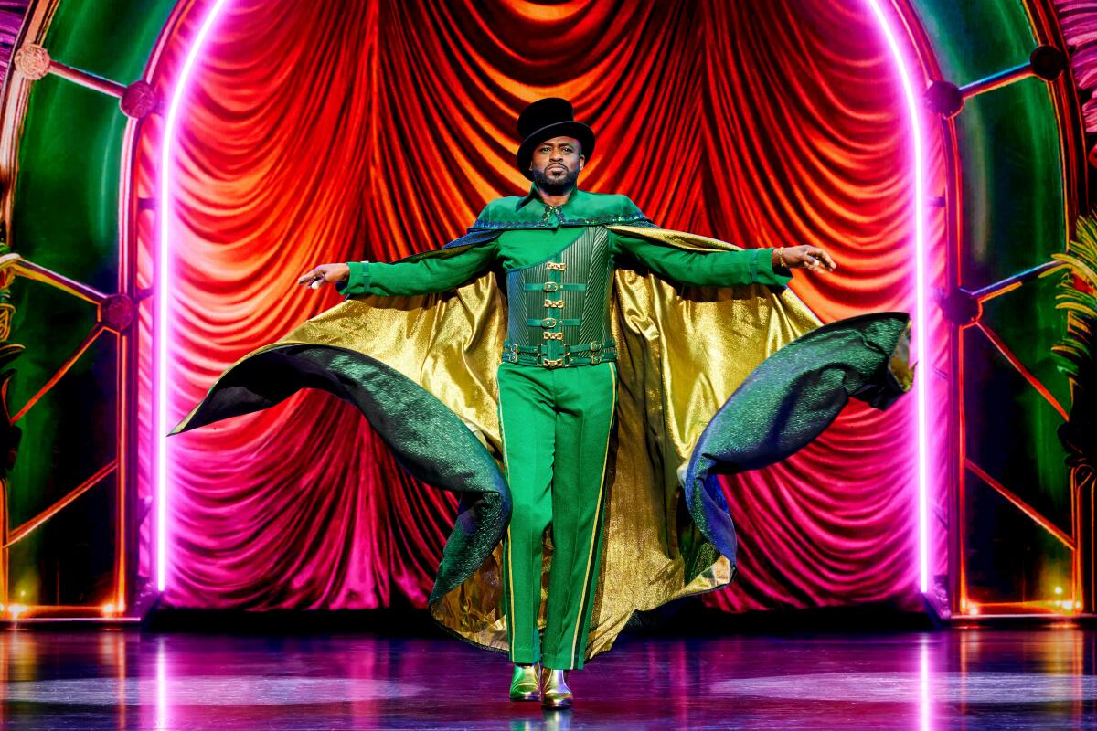 Ein Mann in grünem Outfit und goldgefüttertem grünen Umhang auf der Bühne vor einem roten Vorhang.