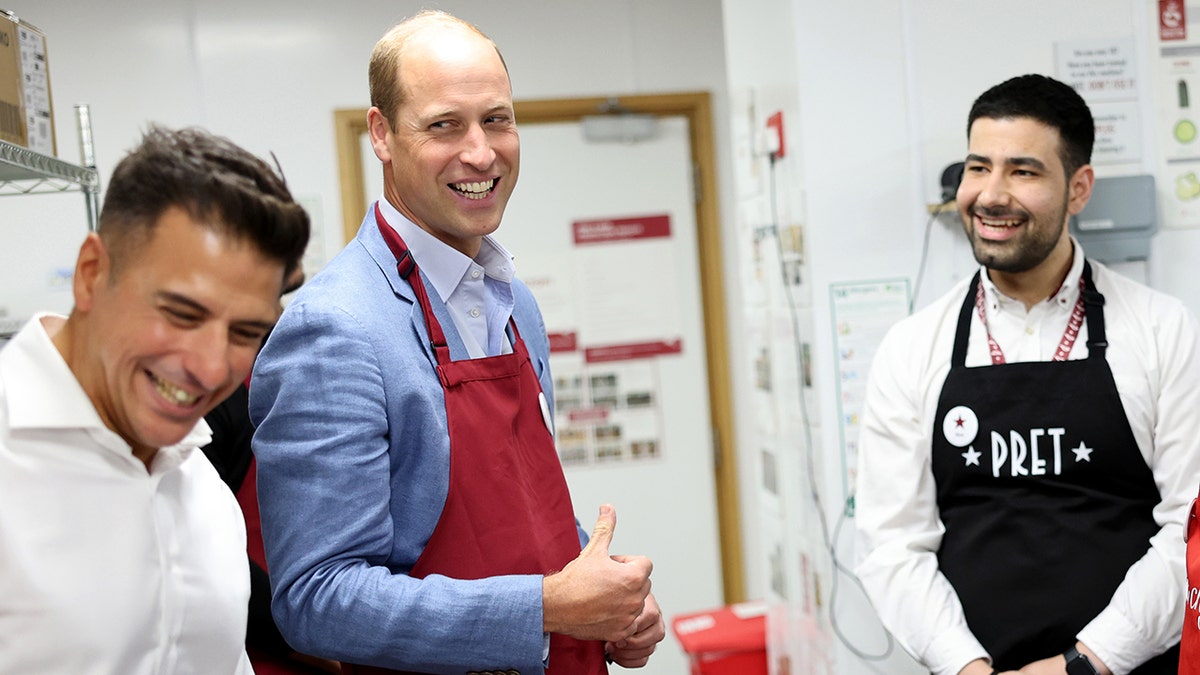 Prinz William in einer Küche, der mit zwei Männern spricht und eine Schürze trägt