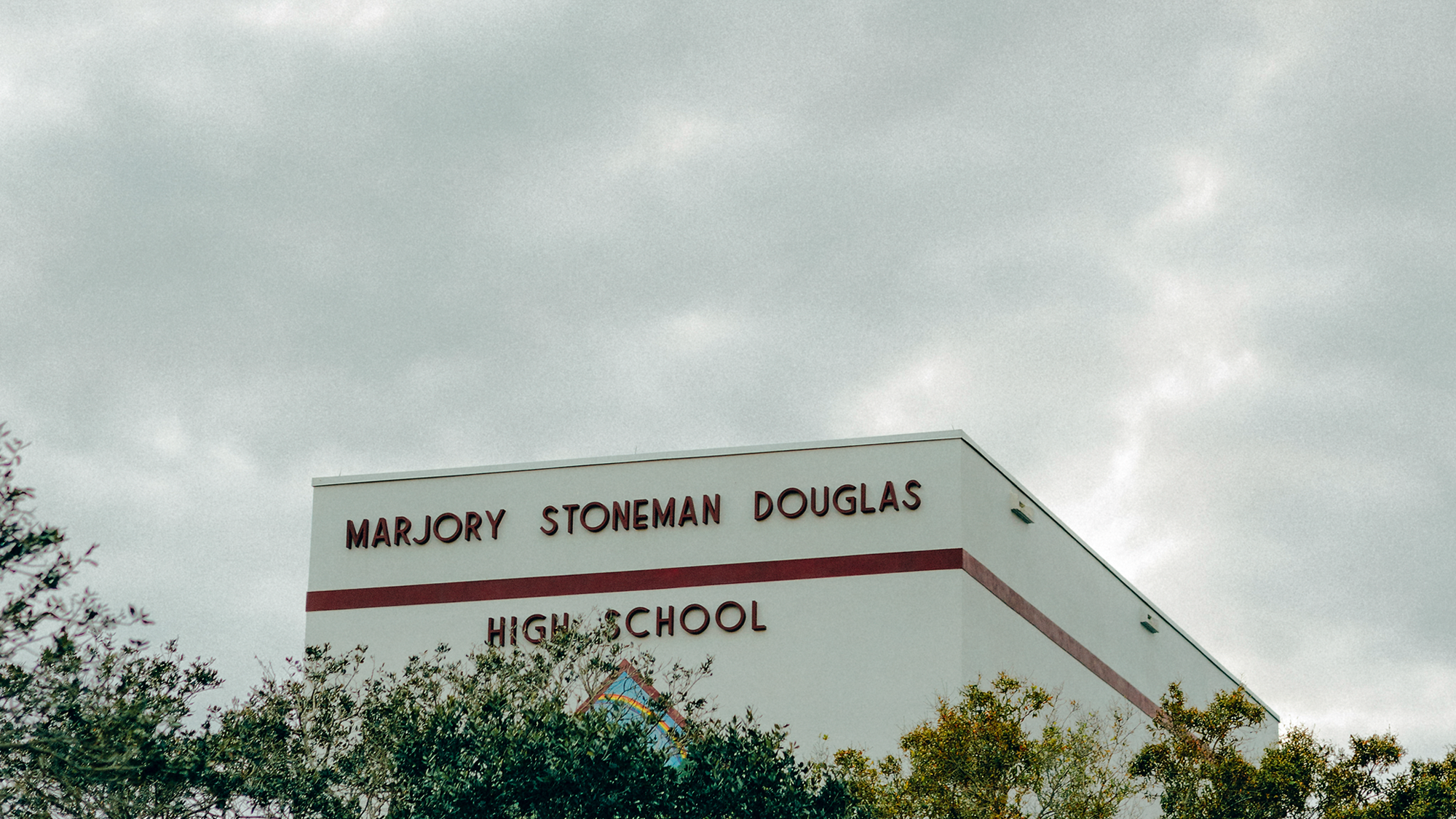 Foto vom Gebäude mit "Marjory Stoneman Douglas High School" darauf, hinter Bäumen vor wolkigem grauen Himmel