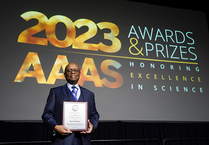 Sekazi Mtingwa hält eine Tafel vor einen Bildschirm, auf der steht: "2023 AAAS Awards & Preise zur Würdigung herausragender wissenschaftlicher Leistungen"