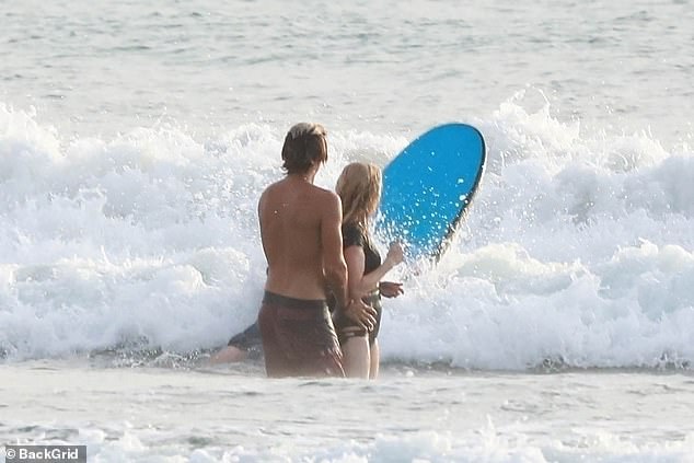 Armando legt seine Hände auf Ellies Taille, während sie einem anderen Strandbesucher beim Surfen zusehen