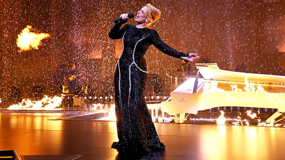 Adele lehnt sich in einem schwarzen Kleid zurück und singt auf der Bühne, während im Hintergrund Feuer und Regen zu sehen sind