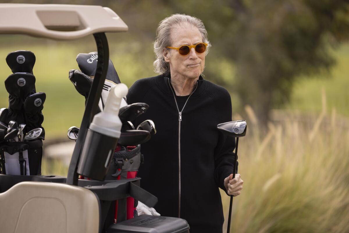 Richard Lewis, ganz in Schwarz gekleidet, hält einen Golfschläger in der Hand und steht neben einem Golfwagen. 