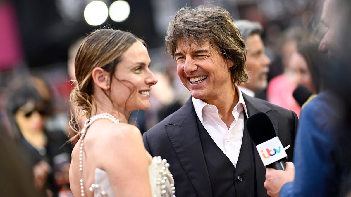 Tom Cruise im schwarzen Anzug blickt Rebecca Ferguson auf dem Teppich sehnsüchtig an, während sie interviewt werden