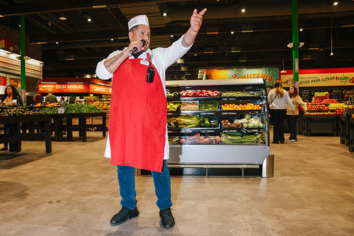 Ein Mann in einer roten Schürze, der ein Mikrofon in der Hand hält, steht mitten in der Obst- und Gemüseabteilung eines Lebensmittelladens.