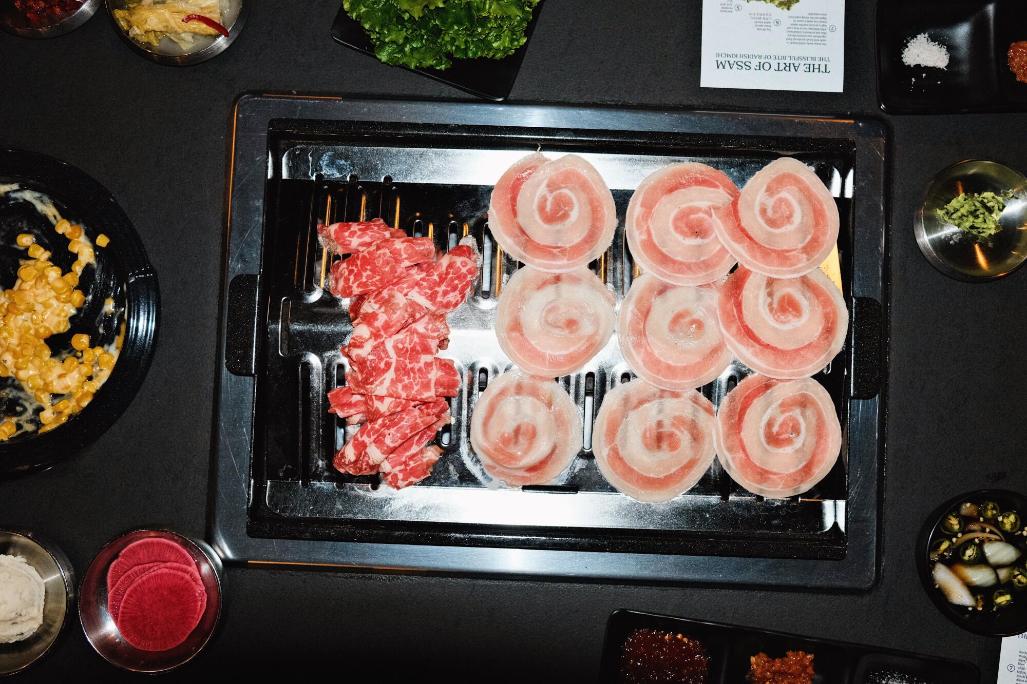 Ein Foto von oben von einem Grill bei Origin Korean BBQ: Die eine Hälfte besteht aus Rinderbrust, die andere aus Schweinebauchscheiben.  Banchan umgeben es.