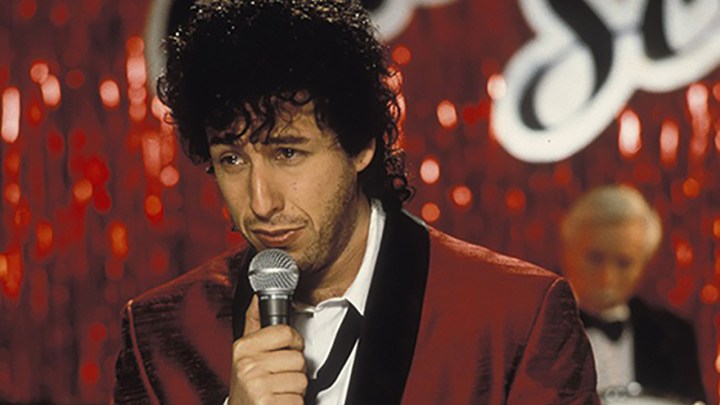Adam Sandler mit lockigem Haar hält ein Mikrofon in der Hand und singt in einer Szene aus The Wedding Singer.