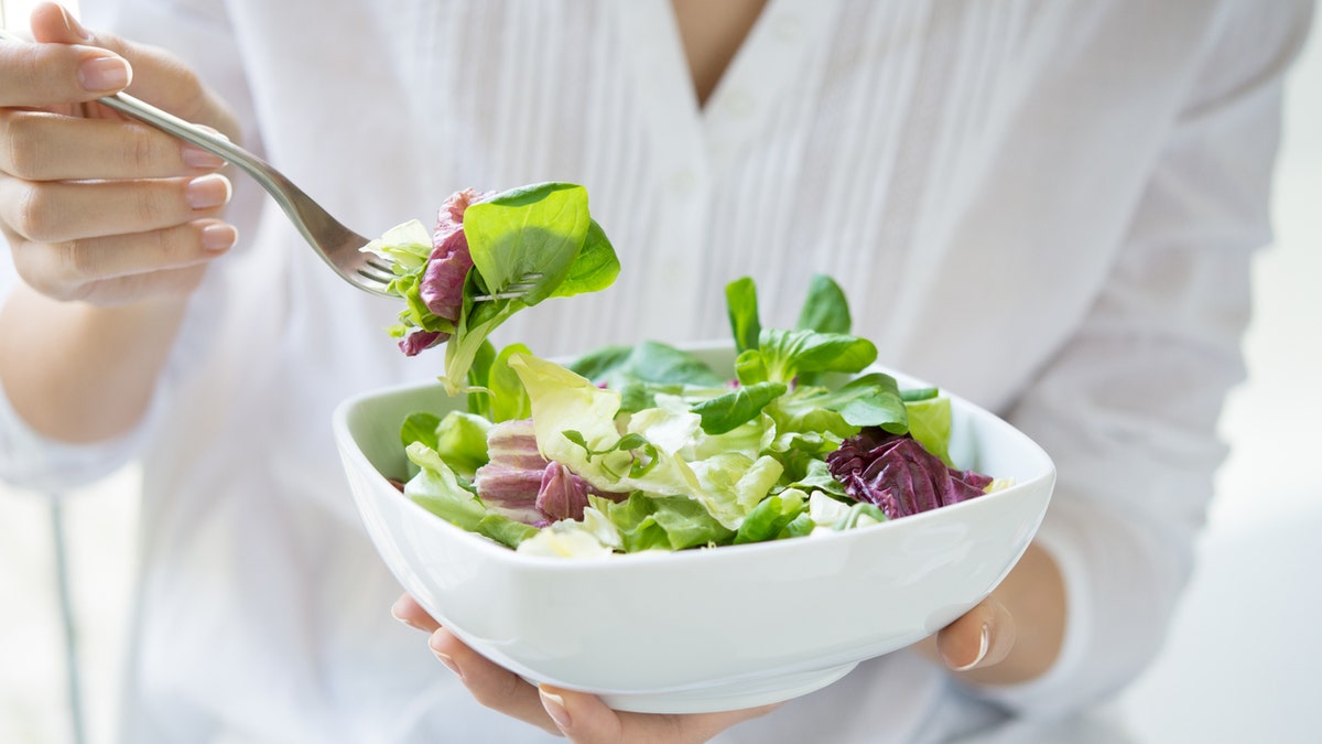 Nahaufnahme einer Frau, die im schönen Morgenlicht einen Teller mit frischem grünen Salat hält.  Sie hält eine Gabel in der Hand und ist dabei, das vegetarische Essen zu essen.  Gesundes Ernährungs- und Diätkonzept.  Geringe Schärfentiefe mit Fokus auf die Gabel.