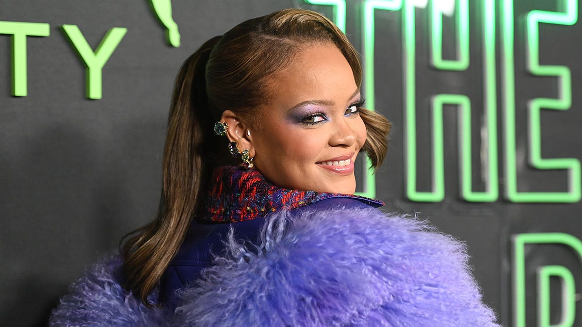 Rihanna schaut in einer zotteligen lila Jacke hinter sich auf den Teppich und lächelt