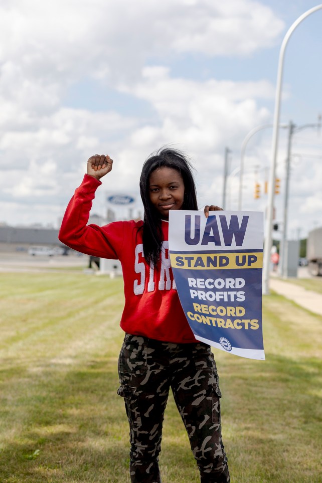Junge Frau im roten Sweatshirt hält mit erhobener Faust ein UAW-Streikplakat in der Hand.