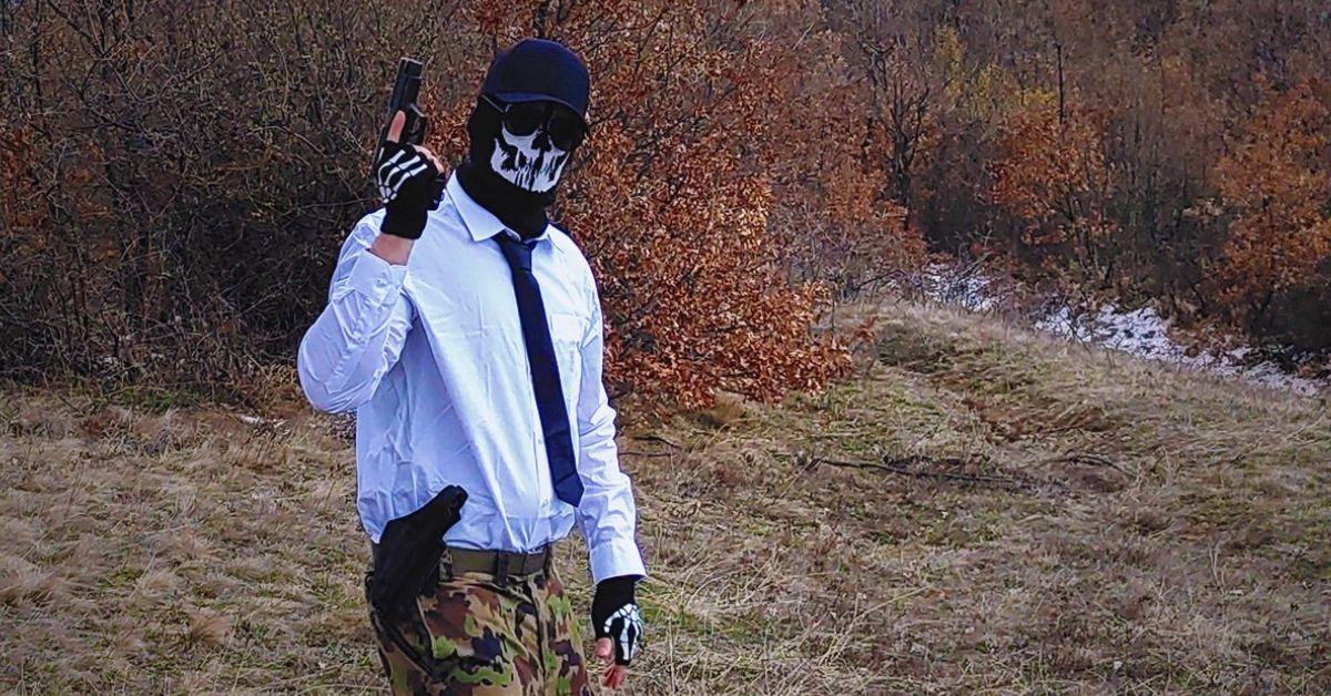 Shadman posiert draußen mit einer Waffe, trägt eine Skelettmaske, Handschuhe, Tarnhosen und ein Button-Down-Hemd mit Krawatte