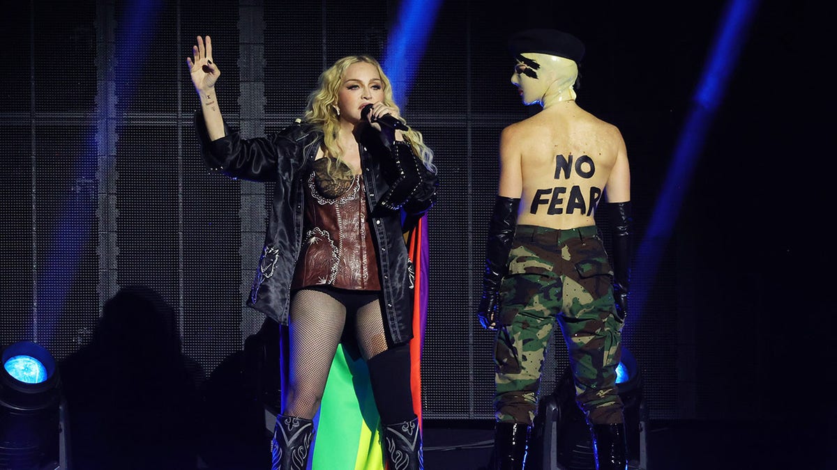 Madonna in einem schwarzen Spitzentrikot, Strumpfhosen und einer dunkelschwarzen Jacke singt auf der Bühne und steht neben einer Tänzerin, auf deren Rücken die Worte „No Fear“ geschrieben stehen