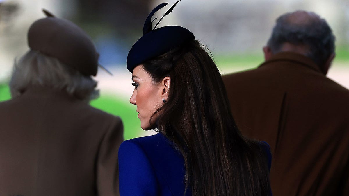 Kate Middleton steht größtenteils mit dem Rücken zur Kamera und trägt einen blauen Mantel und einen Kopfschmuck