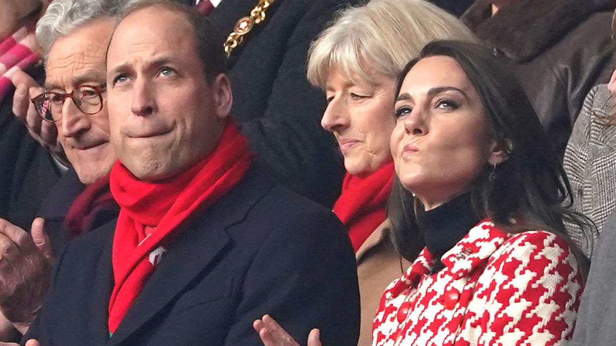 Prinz William in einer dunklen langen Jacke und einem leuchtend roten einfarbigen Schal auf der Tribüne neben seiner Frau Kate Middleton in einer gemusterten rot-weißen Jacke beim Rugbyspiel Wales gegen England