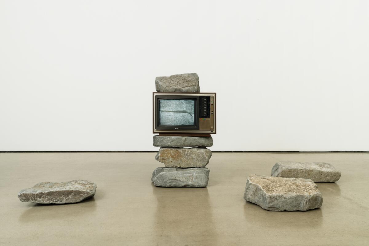 Ein Fernsehmonitor steht auf einem Stapel schwerer Steine, umgeben von weiteren verstreuten Steinen.