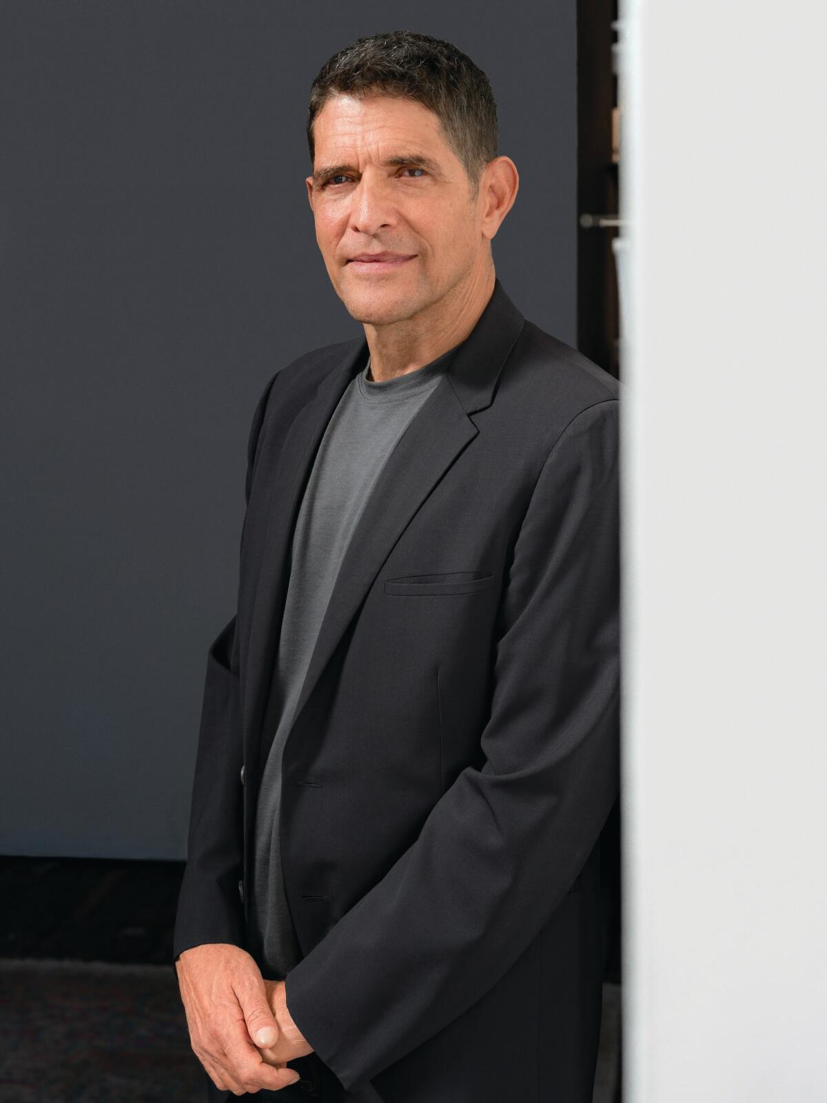 Ein dunkelhaariger Mann in einem dunklen Blazer über einem grauen T-Shirt steht an einer Wand