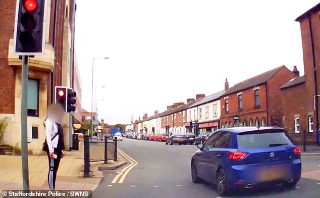 In einem Video ist zu sehen, wie ein rasendes blaues Auto an einer roten Ampel vorbeifährt, gerade als eine Person versucht, die Straße zu überqueren