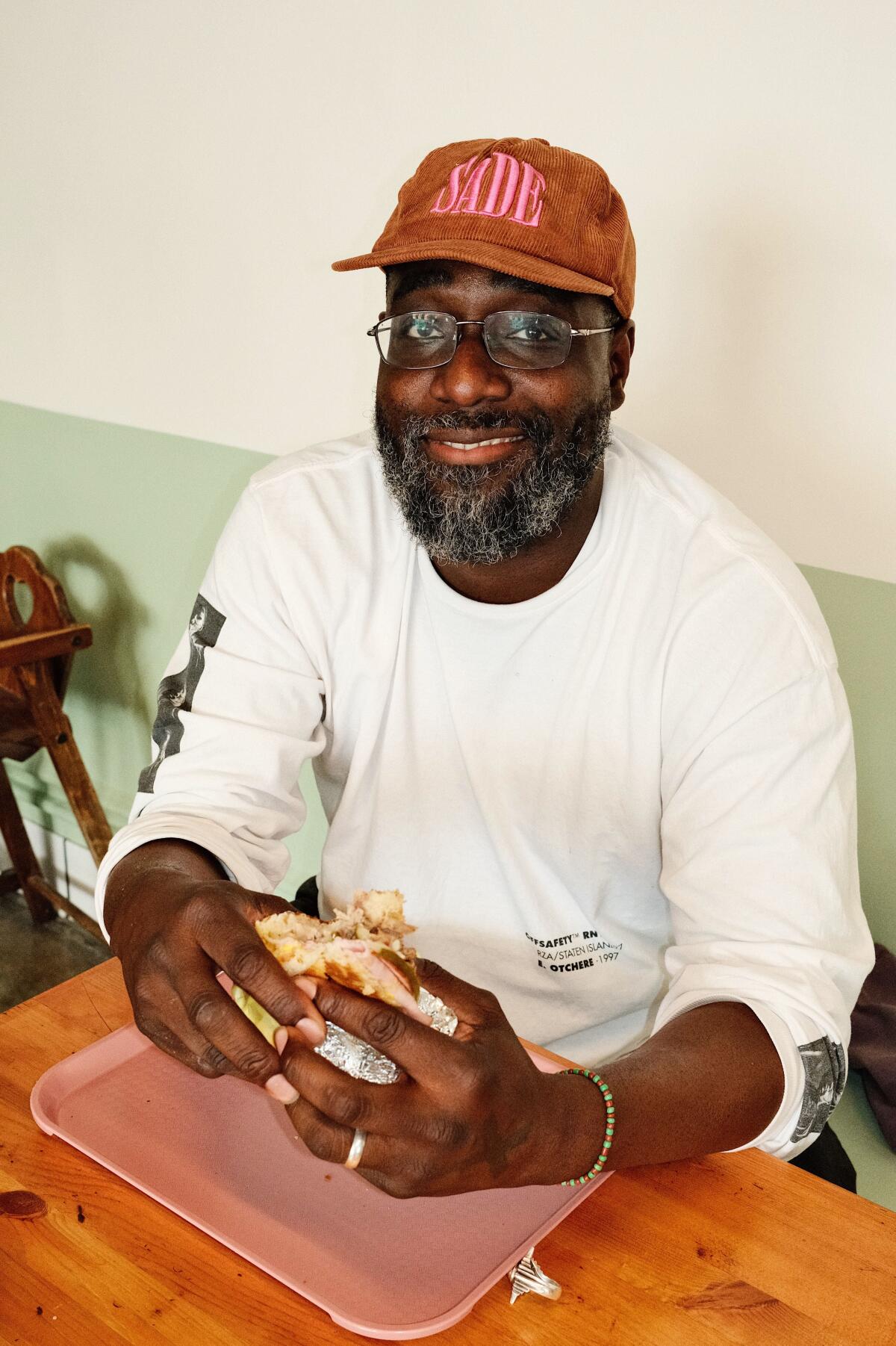 Ein vertikales Foto von Toks Shoyoye, der am Tag der Wiedereröffnung in Silver Lake an einem Stand des Cafe Tropical sitzt und ein Sandwich in der Hand hält